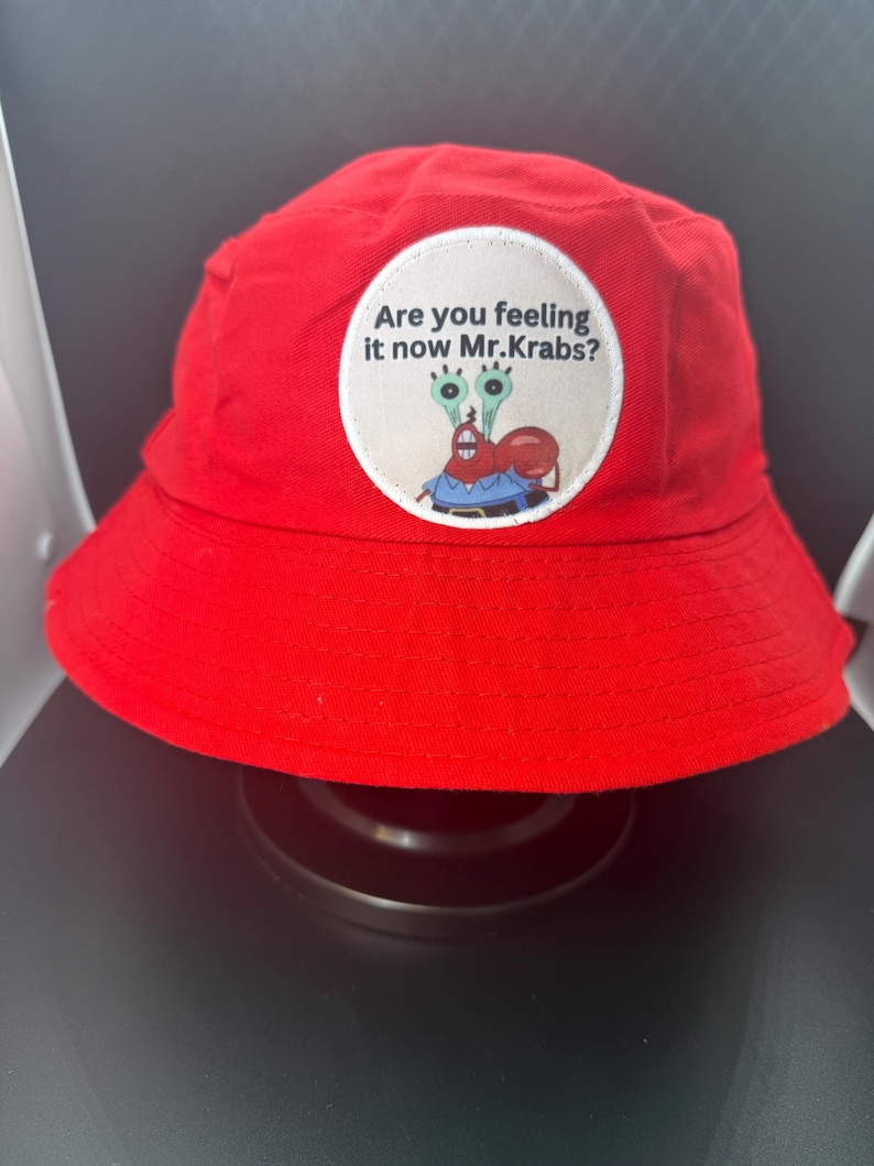 Puede incluir: Sombrero de pescador rojo brillante con un parche circular con Mr. Krabs de Bob Esponja. El parche tiene el texto "Are you feeling it now Mr. Krabs?" en fuente negra. El sombrero est&aacute; hecho de tela suave.