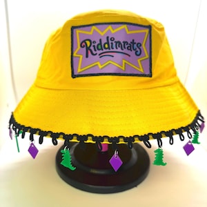 Puede incluir: Un sombrero de pescador amarillo brillante con ribete negro y abalorios colgantes. El sombrero presenta un parche con el texto "Riddinrats" en púrpura y amarillo. Los abalorios incluyen formas de dinosaurios verdes y formas de diamantes púrpuras.