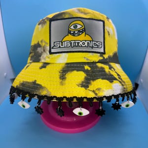 Puede incluir: Sombrero de pescador amarillo teñido anudado con ribete negro y colgantes de ojos. El sombrero presenta un parche con la palabra "SUBTRONICS" y un personaje de dibujos animados con un solo ojo. El sombrero está sobre una superficie rosa.