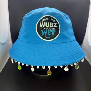 Puede incluir: Sombrero de pescador azul brillante con un gráfico circular que dice "I WANT THE WUBZ TO SOUND WET". El sombrero está adornado con conchas blancas y amuletos en forma de lágrima verdes y amarillos a lo largo del borde.
