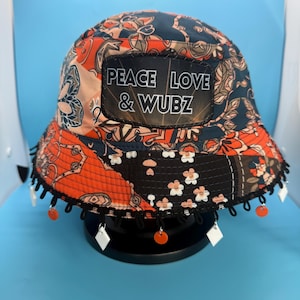 May include: A patterned bucket hat with a black brim and decorative fringe. The hat features a mix of orange, navy, and peach paisley patterns. A patch on the front reads "PEACE LOVE & WUBZ".