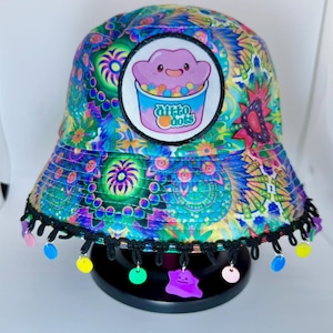 May include: A colorful bucket hat with a vibrant floral and geometric pattern in shades of blue, green, purple, and pink. The hat features a patch with a cartoon ice cream design and the text "Ditto Dots". The brim is trimmed with black fringe and colorful charms.