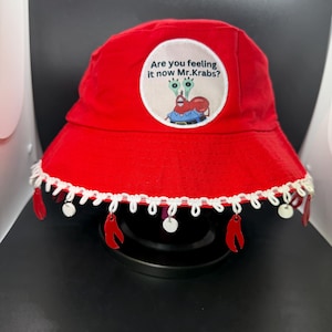 Puede incluir: Sombrero de pescador rojo brillante con ribete blanco y colgantes de garras rojas. El sombrero presenta un parche circular con el texto "Are you feeling it now Mr. Krabs?" y una imagen de dibujos animados de Mr. Krabs.