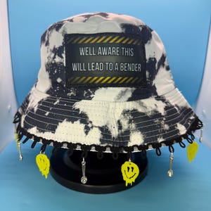 May include: A black and white tie-dye bucket hat with a black lace trim and dangling charms. The hat features a patch with the text "WELL AWARE THIS WILL LEAD TO A BENDER". Yellow smiley face charms and silver spoon charms hang from the brim.