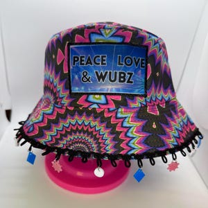 May include: A colorful bucket hat with a psychedelic pattern in pink, blue, and black. The hat features the text "PEACE LOVE & WUBZ" on a rectangular patch. The brim is trimmed with black lace and dangling charms.