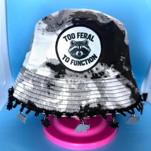 May include: A black and white tie-dye bucket hat with a black lace trim and raccoon charms. The hat features a circular patch with a raccoon graphic and the text "TOO FERAL TO FUNCTION".