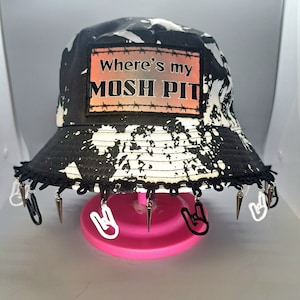 May include: Black and white bucket hat with a white patch that says "Where's my MOSH PIT" and silver spikes around the brim. The hat is on a pink stand with white rock and roll hand signs hanging from it.