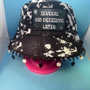 May include: Black and white splatter-print bucket hat with the text "SEVERAL BAD DECISIONS LATER." The hat has a black brim with dangling black and white embellishments. The hat is displayed on a pink stand.