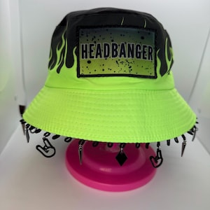 Puede incluir: Un sombrero de pescador verde neón con la parte superior negra y un parche que dice "HEADBANGER". El sombrero presenta diseños de llamas negras y está adornado con colgantes de metal colgantes. El sombrero se muestra sobre un soporte rosa.
