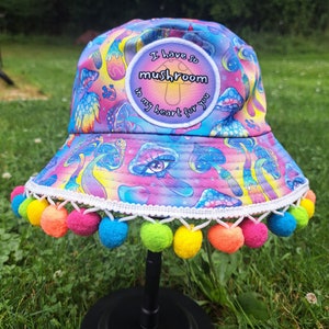 May include: A colorful bucket hat with a mushroom print and a patch that says "I have so mushroom in my heart for you". The hat has a white trim and a row of colorful pom-poms around the brim.