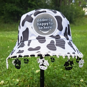 May include: A white and black cow print bucket hat with a white lace trim and cow-shaped charms. The hat has a round patch with the text "Udder-ly happy to be here" in black text.