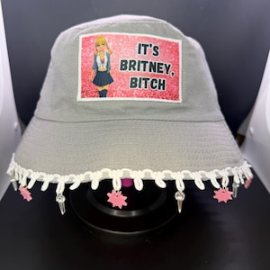 May include: Gray bucket hat with a decorative white lace trim and dangling pink star and crystal charms. The hat features a rectangular patch with the text "IT'S BRITNEY, BITCH" and an illustration of a blonde woman.