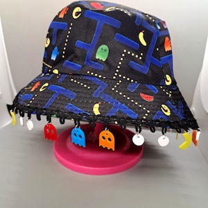 May include: A black bucket hat with a Pac-Man themed design. The hat has a blue and yellow trim and features Pac-Man characters, ghosts, and a maze pattern. The hat has a black lace trim with colorful Pac-Man ghost charms.