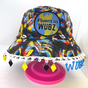 May include: A colorful bucket hat with a vibrant abstract design in blue, yellow, red, and black. The hat features the text "Fluent in WUBZ" on the front and is trimmed with white lace and dangling yellow and black diamond-shaped charms.