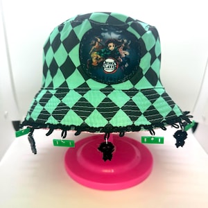 May include: A green and black diamond-patterned bucket hat with a circular patch featuring characters and the text "Demon Slayer". The hat has black trim and dangling charms.
