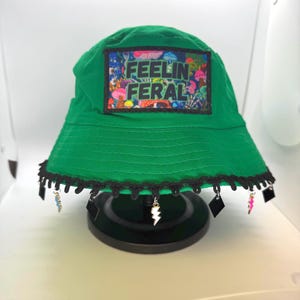 May include: Green bucket hat with a black and white patch that says "Feelin Feral" and a black lace trim. The hat has several charms hanging from the brim, including lightning bolts and squares.