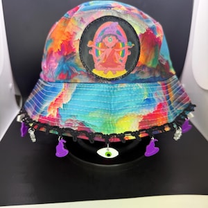 May include: A colorful bucket hat with a vibrant tie-dye pattern in shades of blue, orange, yellow, and pink. The hat features a black patch with a cartoon figure and charms with a witch hat, eye, and silver charms.