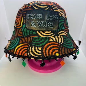 May include: A colorful bucket hat with a repeating pattern of orange, brown, and green swirls. The hat features a patch with the text "PEACE LOVE & WUBZ". The brim is trimmed with black fringe and decorative charms.