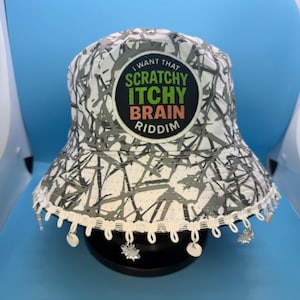 Puede incluir: Un sombrero de pescador con estampado blanco y gris con el texto "I WANT THAT SCRATCHY ITCHY BRAIN RIDDIM" en la parte delantera. El sombrero tiene un ribete blanco con abalorios colgantes. El sombrero está sobre una superficie negra.