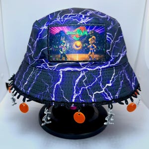 May include: Bucket hat with a blue and black lightning bolt pattern. The hat features a patch with the words "Space Jam" and cartoon characters. The brim is trimmed with black fringe and orange and silver charms.
