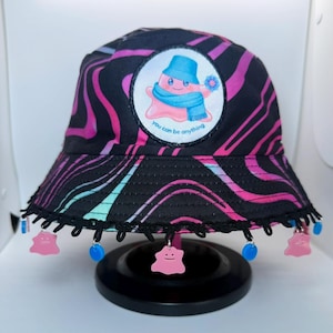 May include: Black bucket hat with a pink and black zebra print. The hat features a patch with a cartoon character and the text "you can be anything." Pink and blue charms dangle from the brim.