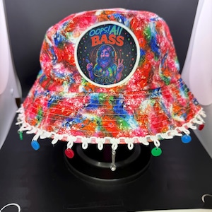 May include: A colorful bucket hat with a vibrant, abstract pattern in red, blue, and green. The hat features a circular patch with the text "OOPS! All BASS" and an illustration of a man with headphones. The brim is trimmed with white lace and colorful dangling accents.