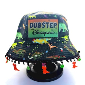 May include: Black bucket hat with a dinosaur print and the text "DUBSTEP Disneyland." The hat has a black trim with orange and green dinosaur charms hanging from it. The hat is on a black stand.