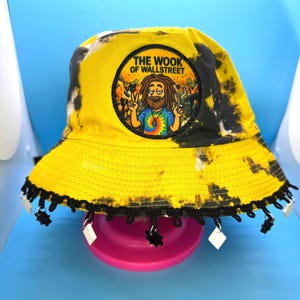 May include: Yellow tie-dye bucket hat with a black trim and dangling charms. The hat features a circular patch with the text "THE WOOK OF WALLSTREET" and a cartoon character. The hat is sitting on a pink surface against a blue background.