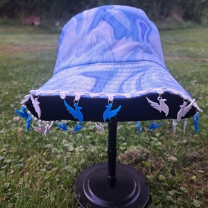 May include: A blue and white tie-dye bucket hat with a black brim and white trim. The hat has blue and white bird charms hanging from the brim.