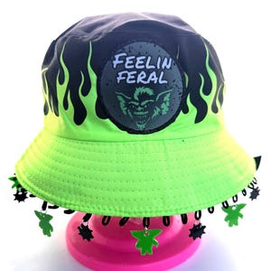May include: A neon green bucket hat with black accents and a patch that reads "FEELIN' FERAL". The hat features a flame design and dangling charms. The hat is displayed on a pink stand.