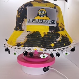 May include: A yellow and black tie-dye bucket hat with a white trim and black stars. The hat has a patch with a cartoon character and the text "Subtronics". The hat is decorated with black stars and green eyes.