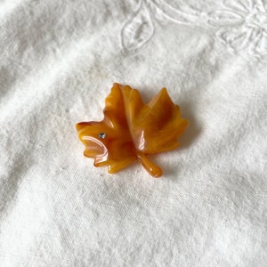 May include: A maple leaf-shaped brooch in shades of amber and brown, with a small, sparkling rhinestone accent. The brooch is made of a glossy material and has a detailed, realistic design. The brooch is a fall-themed accessory.