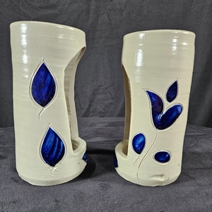 May include: Two white ceramic candle holders with blue floral designs. The holders have a cutout on the side for a candle.