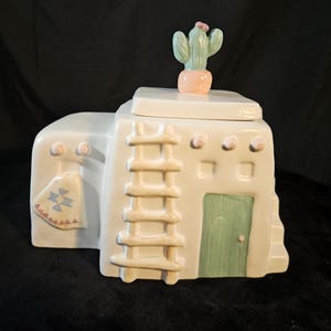 Pode incluir: Jarro de biscoitos de cerâmica branca em forma de casa de adobe do sudoeste com uma escada, uma porta e um cacto no telhado.