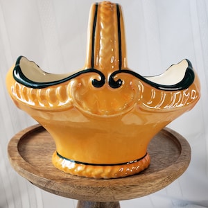 May include: A yellow ceramic basket with a green handle. The basket has a decorative design and is sitting on a wooden stand.