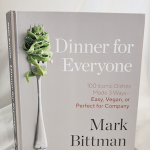 May include: A cookbook titled "Dinner for Everyone" by Mark Bittman. The cover features a fork with a green pasta dish on it. The book promises 100 iconic dishes made 3 ways: easy, vegan, or perfect for company.