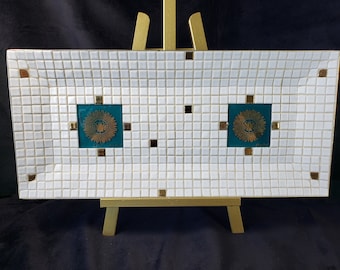 May include: A white ceramic tray with a mosaic tile design. The tray has two gold and teal colored squares with a gold crown design in the center of each square. The tray is on a gold easel.