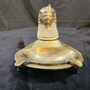 Puede incluir: Un cenicero de latón con una cabeza de faraón esculpida en la parte superior. El cenicero tiene forma ovalada con un borde elevado y una pequeña hendidura en el centro.