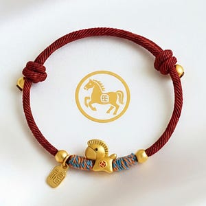 Peut inclure: Bracelet avec un charm en forme de cheval doré et un cordon rouge. Le bracelet comprend un charm en forme de cheval, des perles dorées et une petite étiquette. Un emblème de cheval doré est également visible.