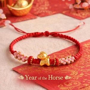 May include: A red braided bracelet with gold details and a gold horse charm. The bracelet has pink floral accents and is on a red card with the text "Year of the Horse".