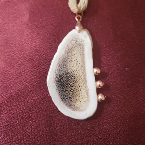 May include: A pendant necklace featuring a white, teardrop-shaped pendant with a brown and black speckled design. The pendant is accented with copper-colored beads and a matching bail, suspended from a beige cord.