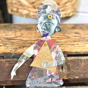 May include: A mixed-media art piece featuring a stylized figure with a gray face and a patterned headdress. The figure has a gold triangle on the chest and a purple dragonfly on the face. The artwork is made of various patterned papers.