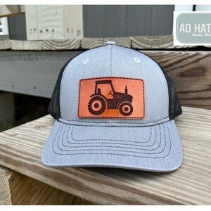 May include: A gray and black trucker hat with a brown leather patch featuring a black silhouette of a tractor. The patch has the text "AO HAT CO.  Clayton, Alabama" on the hat.