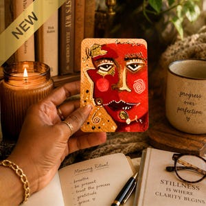 Puede incluir: Una tarjeta de arte rectangular que presenta un rostro abstracto pintado en tonos vibrantes de rojo, oro y negro. La obra incluye rasgos faciales estilizados, patrones en espiral y acentos de texto manuscrito sobre una superficie de papel texturizado.