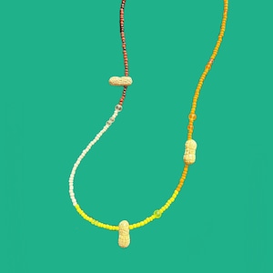 May include: A unique necklace featuring a peanut pendant and colorful beaded strands. The necklace has a split design with red, white, and yellow beads on one side and orange and yellow beads on the other. The necklace is a fun and quirky accessory.