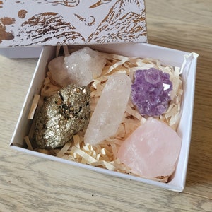 Puede incluir: Una caja blanca con un diseño dorado en la tapa, que contiene una colección de cristales. Los cristales incluyen cuarzo transparente, amatista, cuarzo rosa y pirita.