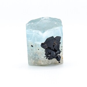 May include: A light blue aquamarine crystal with black inclusions. The crystal is faceted and has a hexagonal shape.
