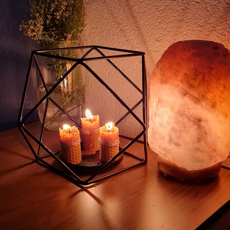May include: Three small, honey-colored candles are lit inside a black metal cube-shaped holder. A large, pink Himalayan salt lamp is on a wooden base to the right of the holder.