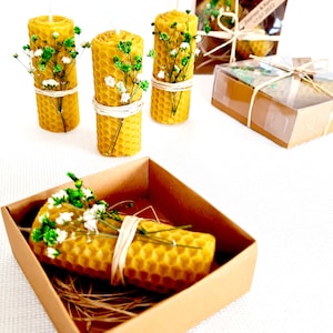 May include: Three small, yellow honeycomb candles with white flowers and twine wrapped around them. The candles are in a small, brown box with shredded paper inside. The box is on a white surface.