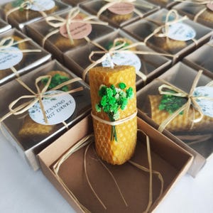 Peut inclure: Bougies en cire d'abeille faites à la main, avec une texture en nid d'abeille et ornées d'un petit bouquet de fleurs vertes. Les bougies sont présentées dans des boîtes transparentes nouées avec du raphia, créant une esthétique rustique et élégante.